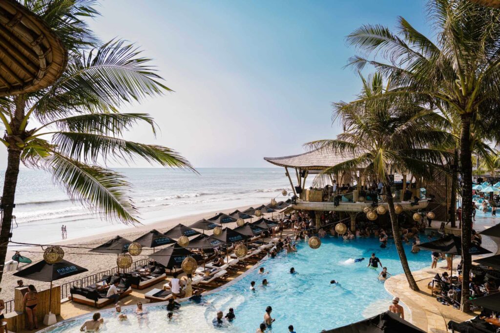 Beachfront pool with sun loungers, black umbrellas and palm trees overlooking the ocean at a Bali beach club