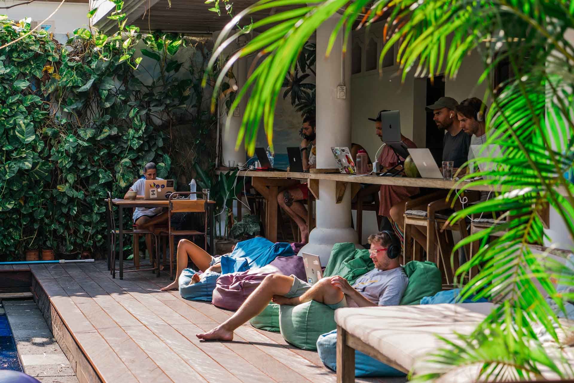 Remote workers on laptops at an open-air Bali coworking cafe surrounded by tropical plants and beanbag seating by a pool
