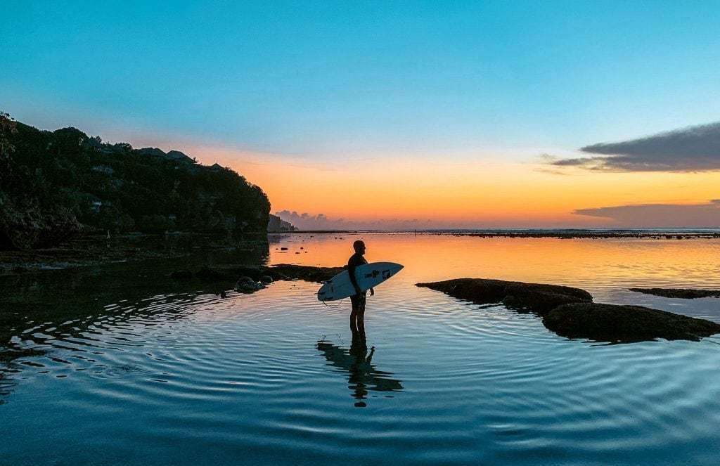 Silhouette of a surfer holding a board at a glassy Bali reef break as the sky glows orange at sunset
